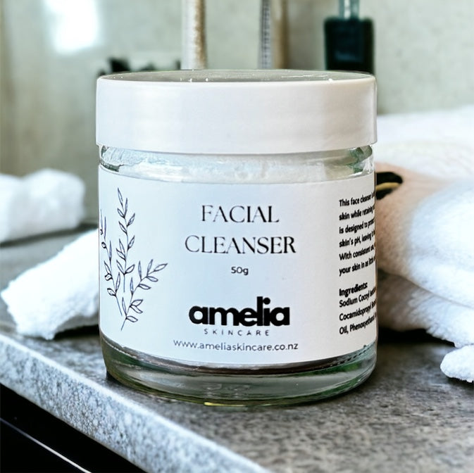 A clear jar with a white lid labeled 'FACIAL CLEANSER 50g' from amelia skincare, containing a white foam, placed on a bathroom countertop.